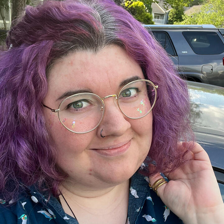 A picture of a person with purple hair and gold glasses standing outside