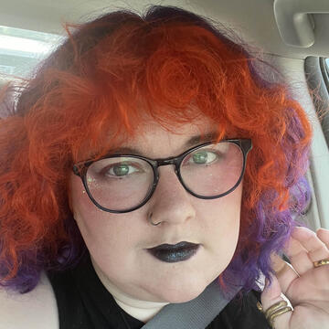 A picture of a person with purple hair and gold glasses standing outside